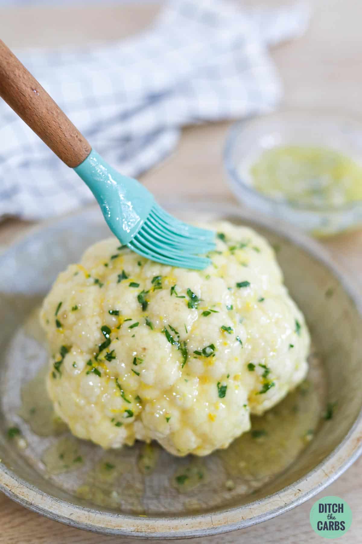 a blue pastry brush brushing melted herbed butter onto a whole cauliflower