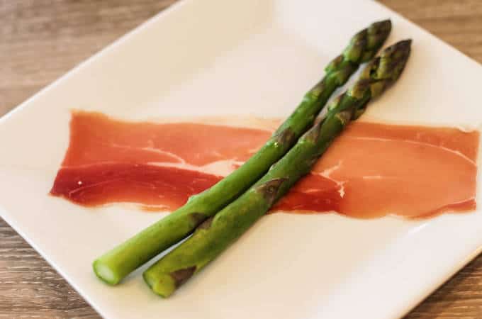 A piece of broccoli on a plate, with Prosciutto wrapped asparagus