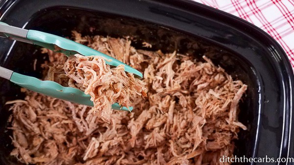 Blue tongs lifting keto pulled pork from the slow cooker 