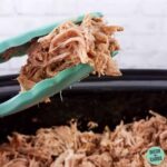 pulled pork being lifted from the slow cooker with blue tongs