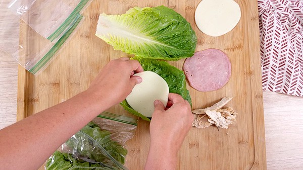 A wooden cutting board with ham and cheese on lettuce leaves being rolled as a wrap