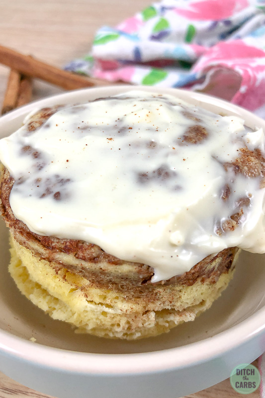 Frosted cinnamon roll low-carb mug cake in a light blue dish with cinnamon sticks on the side.