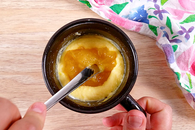 The back of a fork is pushing the lemon curd filling into the cake batter in a black mug.