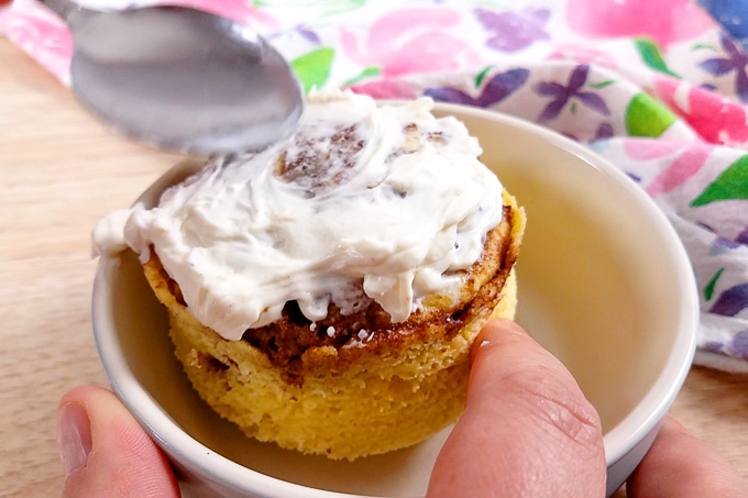 Putting cream cheese icing on a cooked mug cake in a light blue dish with a spoon.