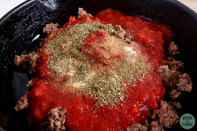 Making meat sauce for low-carb skillet lasagna. The ground beef has been browned in a cast iron pan. Crushed tomatoes has been added on top with seasonings to make the meat sauce.