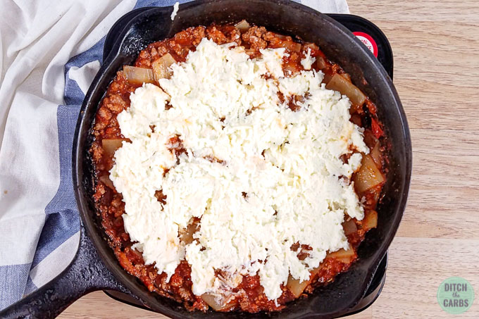Low-carb skillet lasagna ready to bake in a cast iron skillet. The cheese mixture has been spread over the top of the lasagna and the noodle have been mixed into the meat sauce.