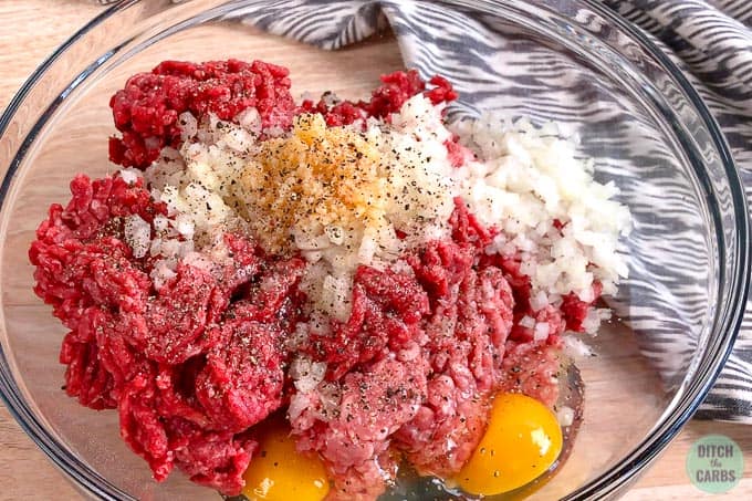 Uncooked ground beef and ground pork in a glass bowl with chopped onions, garlic, seasoning, and two eggs.