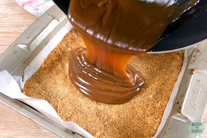 Pouring the caramel for keto caramel slices over the baked almond flour crust in a square pan.