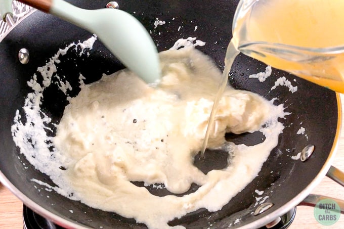 Pouring chicken broth in a glass measuring cup into the melted goat cheese in a skillet.
