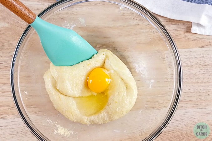 Mixing an egg into the Fat Head dough in a clear glass bowl with a light green spatula.