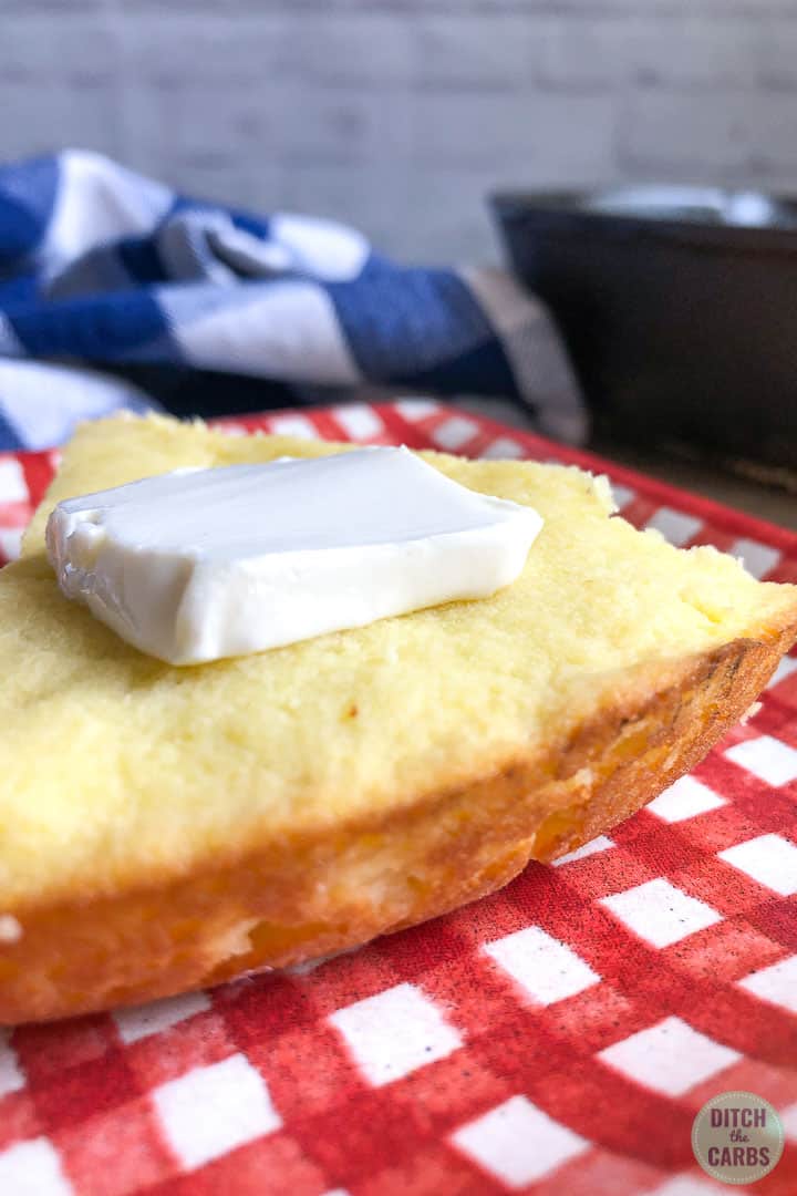 A slice of keto cornbread on a red checkered plate with a slice of butter on top. The crust of the cornbread is facing towards you to show the crisp golden edges.