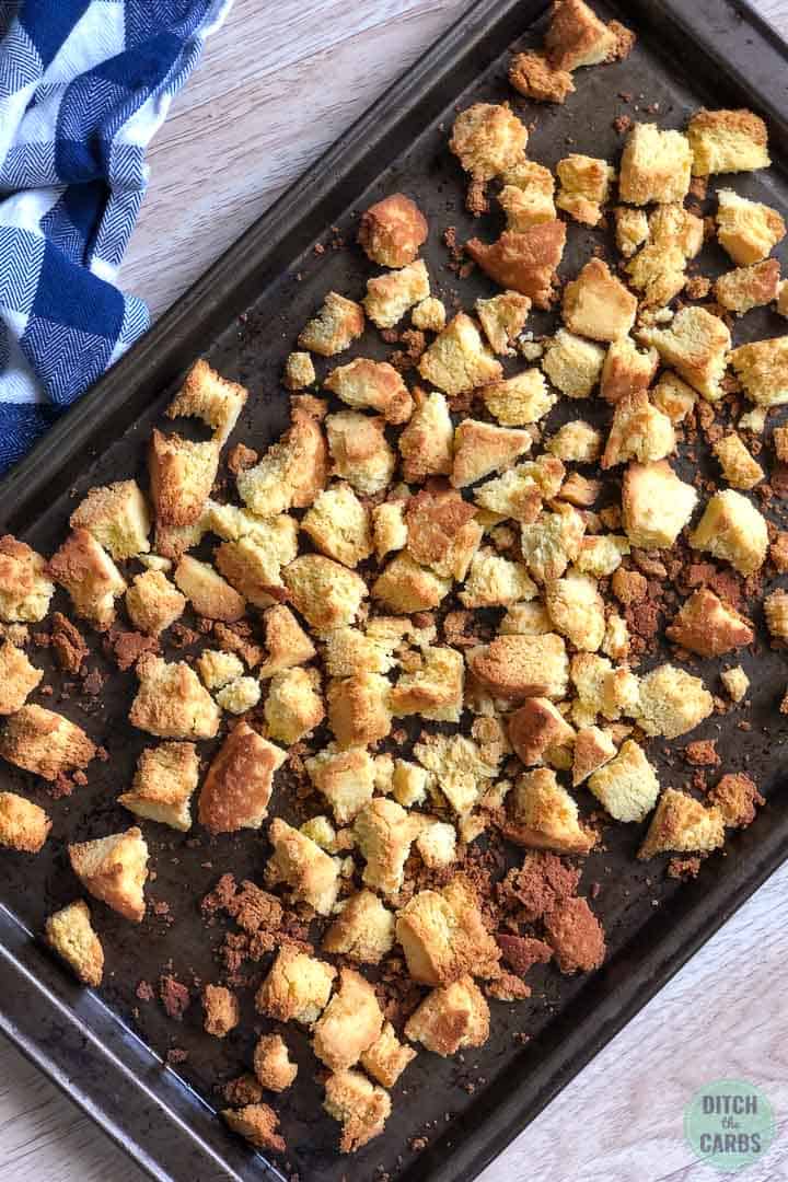 Keto cornbread is broken into pieces and spread out on a baking sheet. The corn have been dried in the oven so that it can be turned into bread crumbs.
