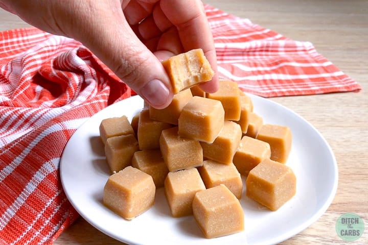 Peanut butter fudge stacked in a pile on a white plate. A hand is picking up a fudge square with a bite taken out of it.