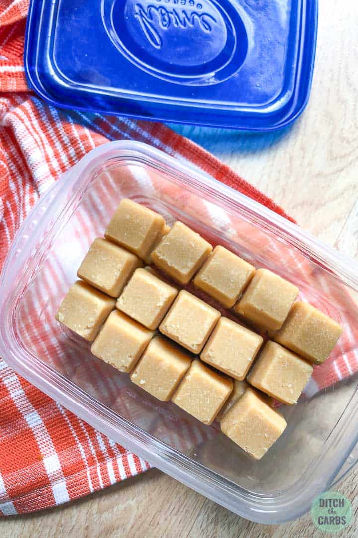 Peanut butter fudge in a small plastic storage container with a blue lid. The fudge is stacked in rows on top of each other.