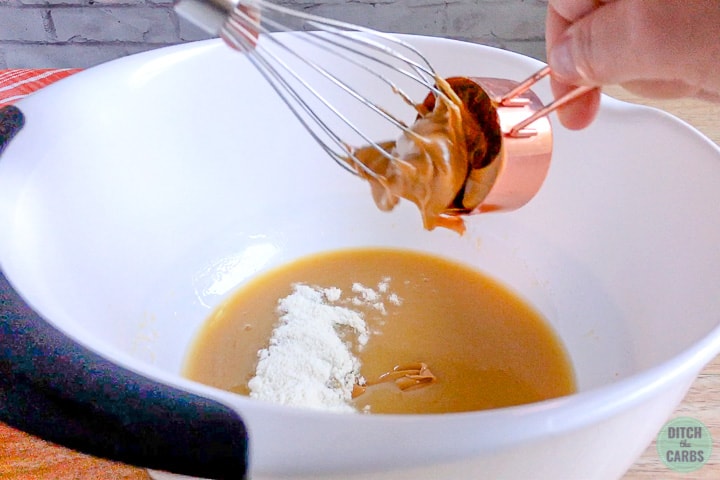 A whisk is used to mix in coconut flour and peanut butter into the condensed keto peanut butter fudge mixture in a large white bowl.