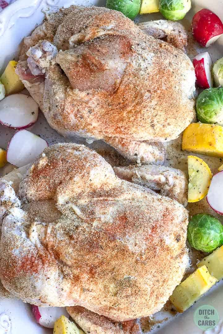 A close up of two Cornish hens smother with butter and seasoned in a white baking pan. Chopping vegetables surround the hens in the pan.