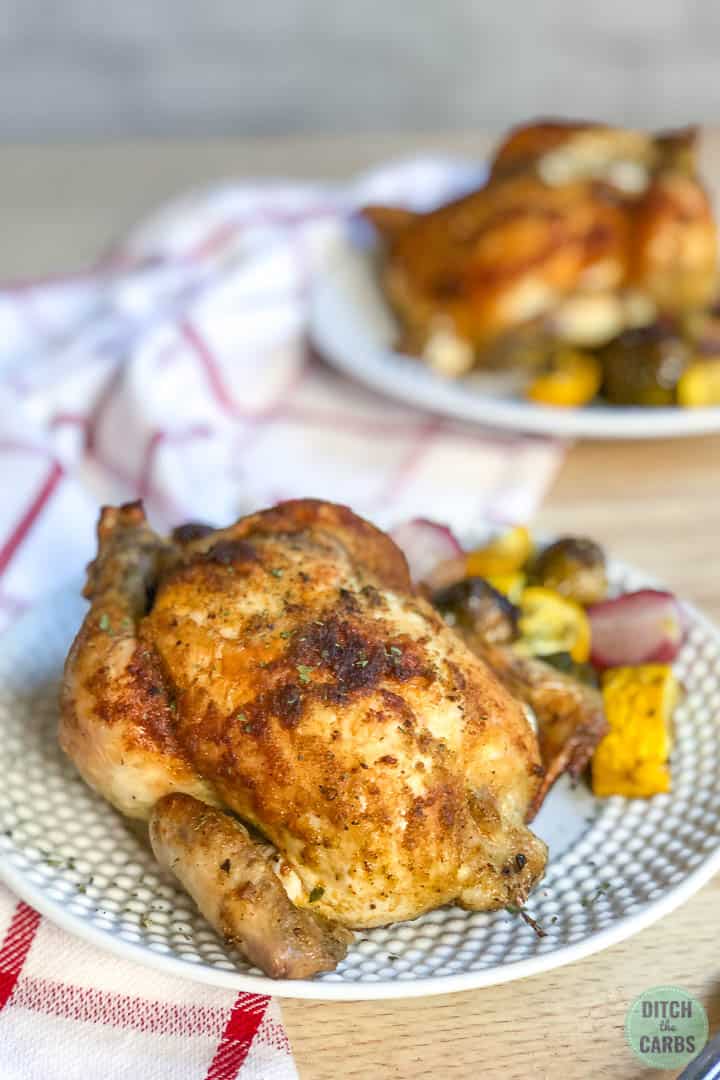 One keto crispy Cornish hen on a white place with roasted vegetables on the side. Another plate with a hen and vegetables is in the background.