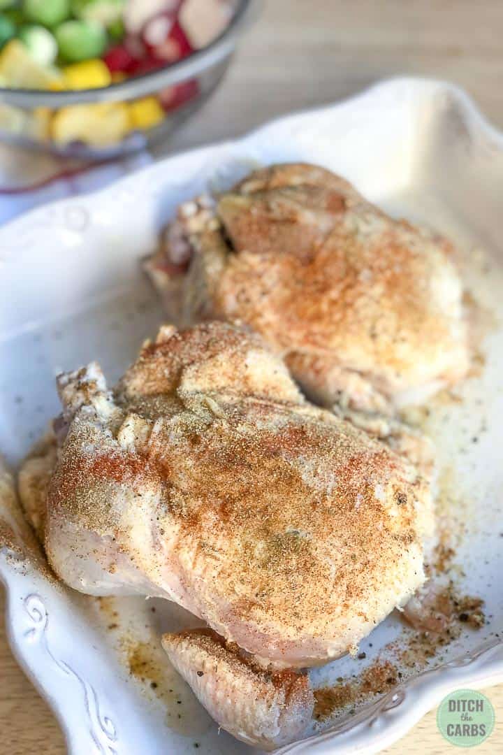 Seasoned  uncooked Cornish hens in a white baking pan. Chopped vegetable are in a clear glass bowl in the background.