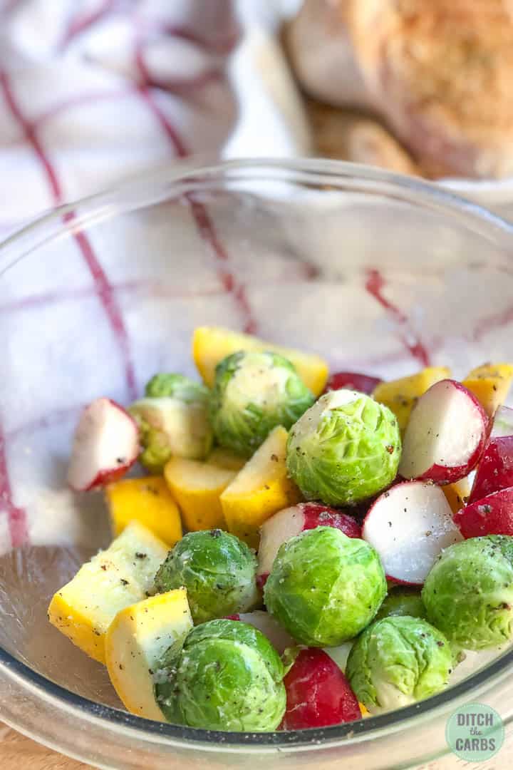 Brussels sprouts, yellow squash, and red radishes tossed together in a clear glass mixing bowl with melted butter, salt, and pepper.