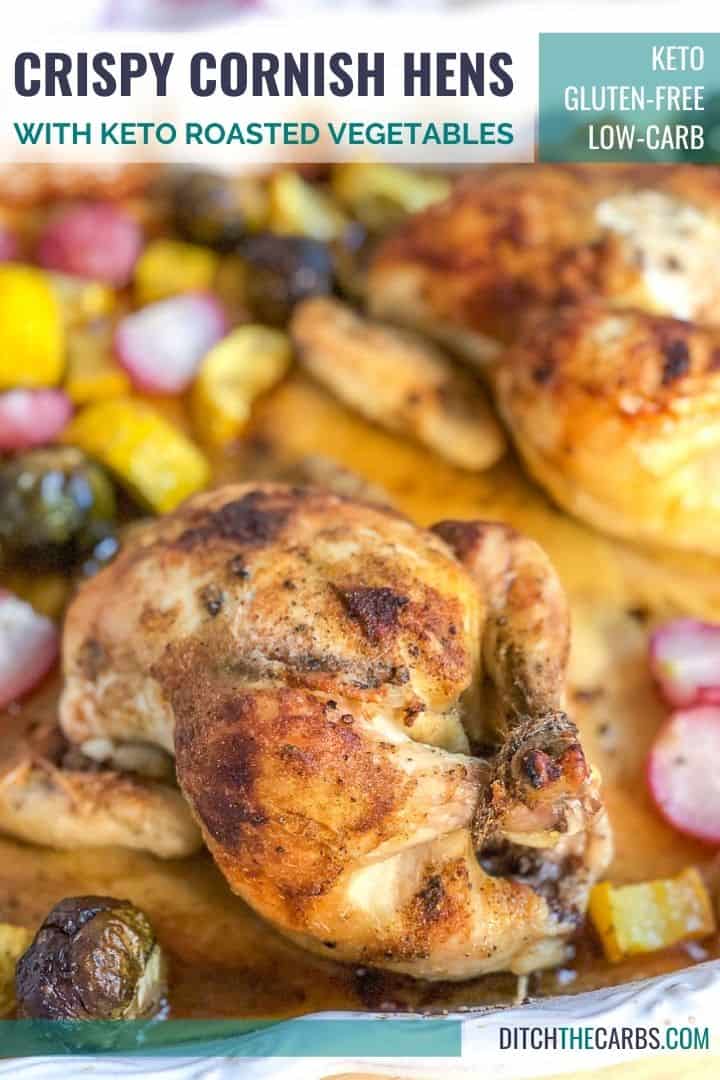 Cooked Keto Crispy Cornish Hens in a white baking pan surrounded by low-carb roasted vegetables. The legs of the hens are tied together with twine.