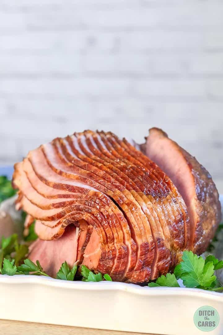 Cooked keto ham in a white baking dish with green parsley underneath a piece of ham as an ornament.  The ham is sliced ​​and ready to serve.
