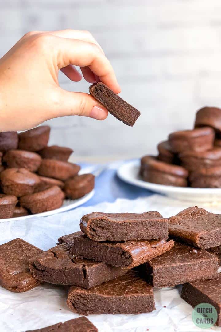 A hand is picking up a dairy-free brownie from a plate fill of square brownie slices. There are white plates full of  brownie muffins in the background.