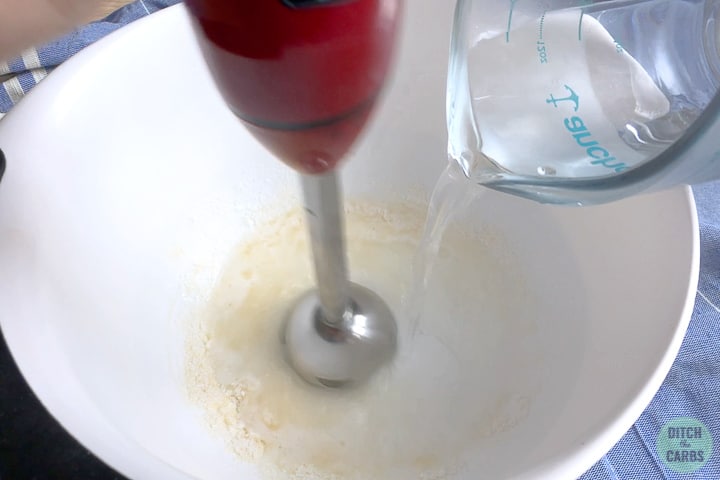 Pouring boiling water into the mixing bowl with gelatin and sweetener. The water is being mixed with an electric stick blender.