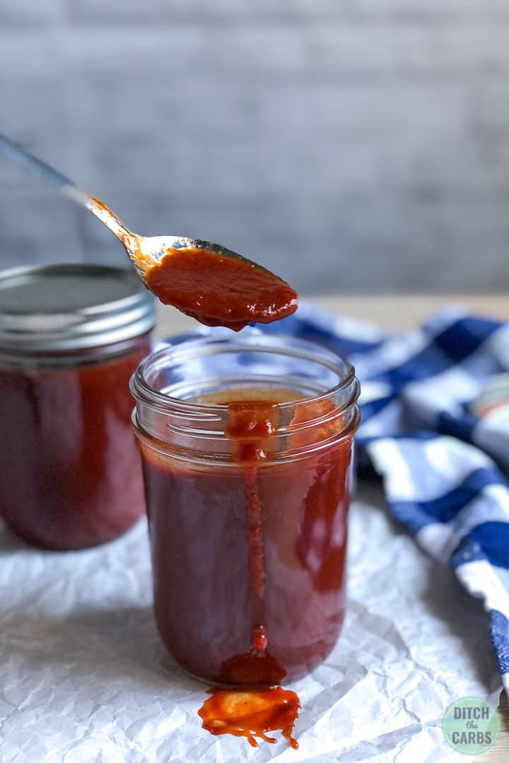 A spoonful of keto BBQ sauce is being lifted from a jar of BBQ sauce. A small drizzle of BBQ sauce is dripping over the edge of the jar.