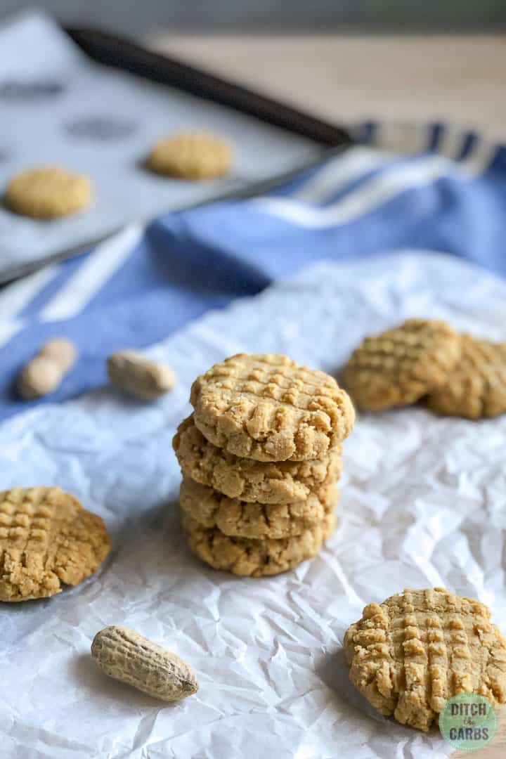 4 keto cooked peanut butter stacked on a white parchment.  More biscuits are laid out beside them and whole peanuts are scattered around the background.