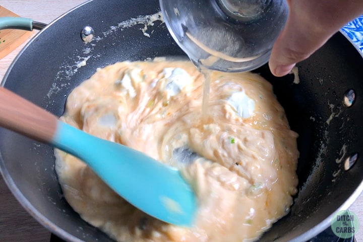 A rubber spatula is slowly stirring chicken broth into the keto cheese sauce in a deep skillet.