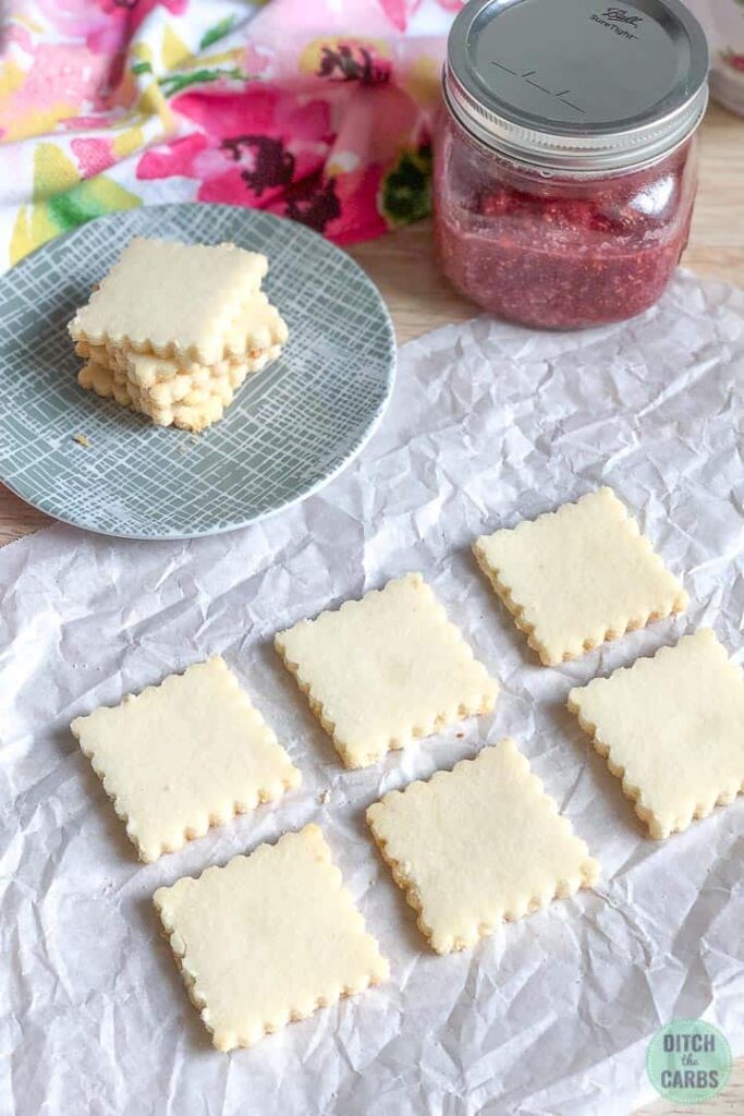 Six keto shortbread cookies rest on a sheet of white parchment paper. The rest of the cookies are stacked on light green stripped dish to the size. Red raspberry jam in a jar sits to the right of the plate.