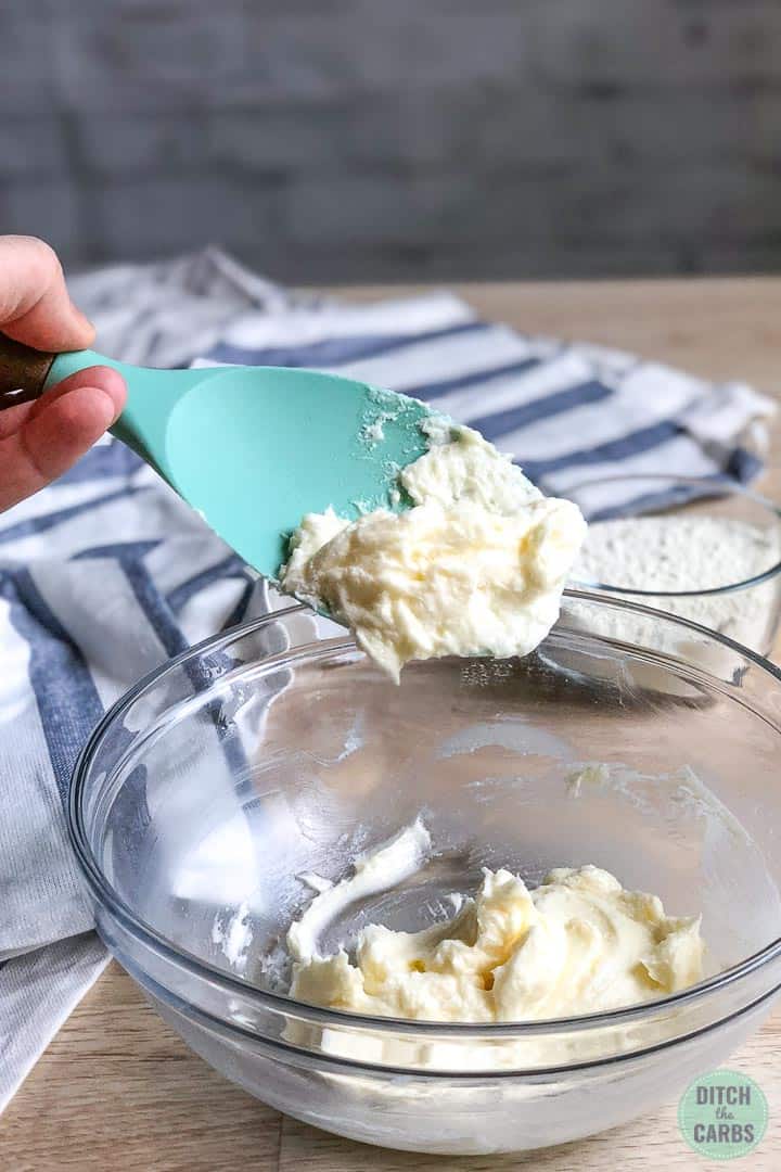 Softened butter is mixed with sweetener in a glass  bowl. A light green rubber spatula is lifting some of the butter mixture out of the bowl.