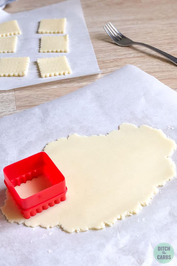 La masa de galletas de mantequilla cetogénica se extiende sobre un trozo de papel pergamino blanco.  Un cortador de galletas con un cuadrado rojo corta las galletas.