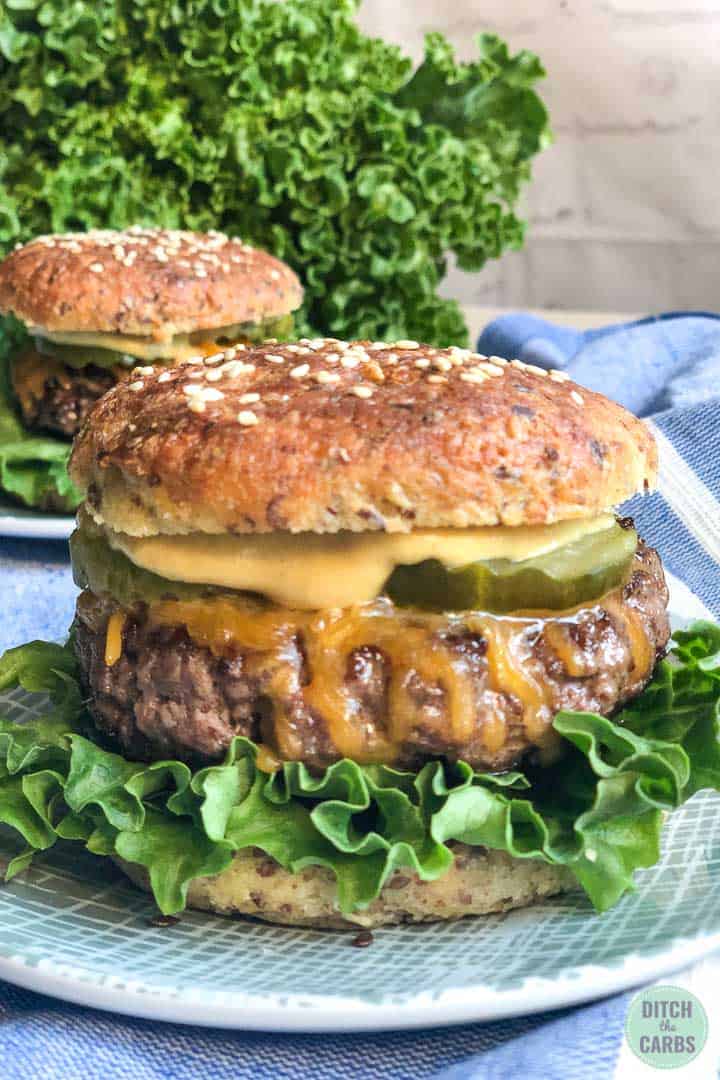 Close of shot of keto hamburger buns on a plate. The bun has been cut a patty, mustard, lettuce, and pickles is inside.