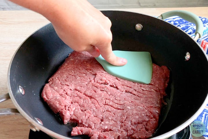 A blue green spatula is pressing the ground beef flat into the bottom of a black skillet to cook.