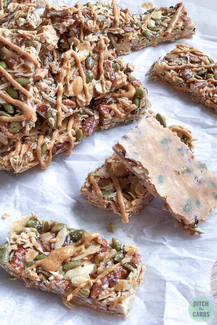 Keto granola bars resting on white parchment paper. One granola bar is turned over to show the bottom of the bar is coated in a layer of peanut butter. The top of the bar is drizzled with peanut butter.