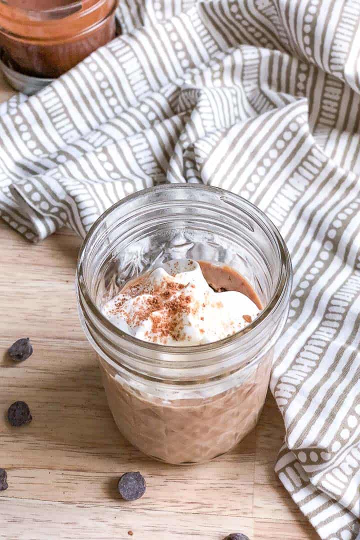 "Nutella" keto mug cheesecake in a clear glass jar resting on a light wood counter. It is topped with sweetened whipped cream and cocoa powder.