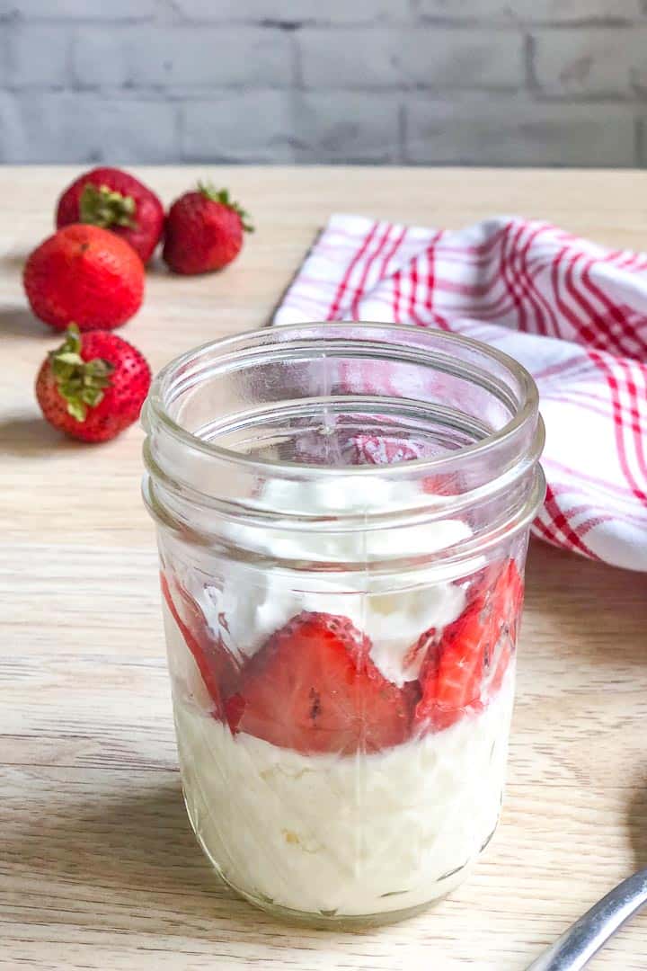 Strawberry keto mug cheesecake cooked cooked in a clear glass jar and topped with fresh strawberries and whipped cream.