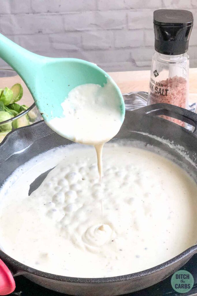 A light green spoon is dripping cheese sauce back into the cast-iron skillet to show how creamy the sauce is.