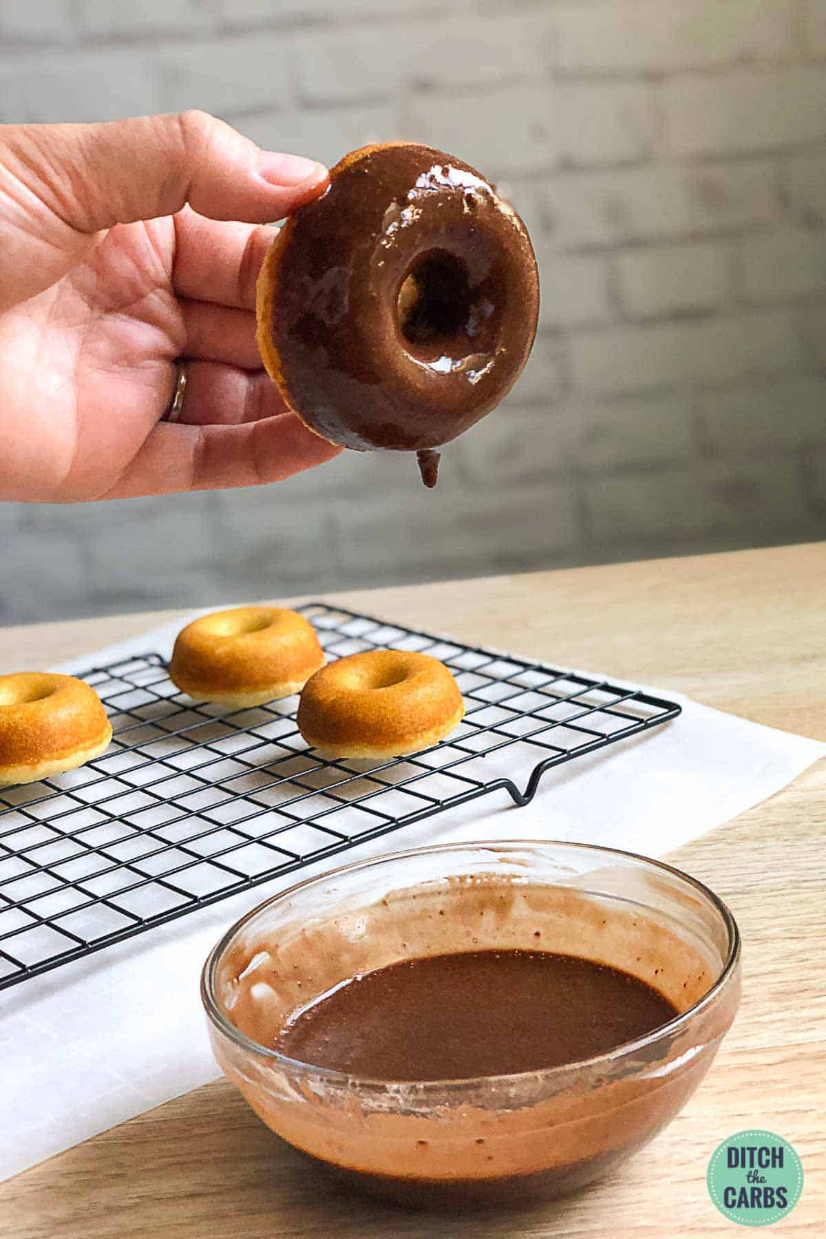 A hand is dipping an almond flour keto donut in a bowl of chocolate icing.