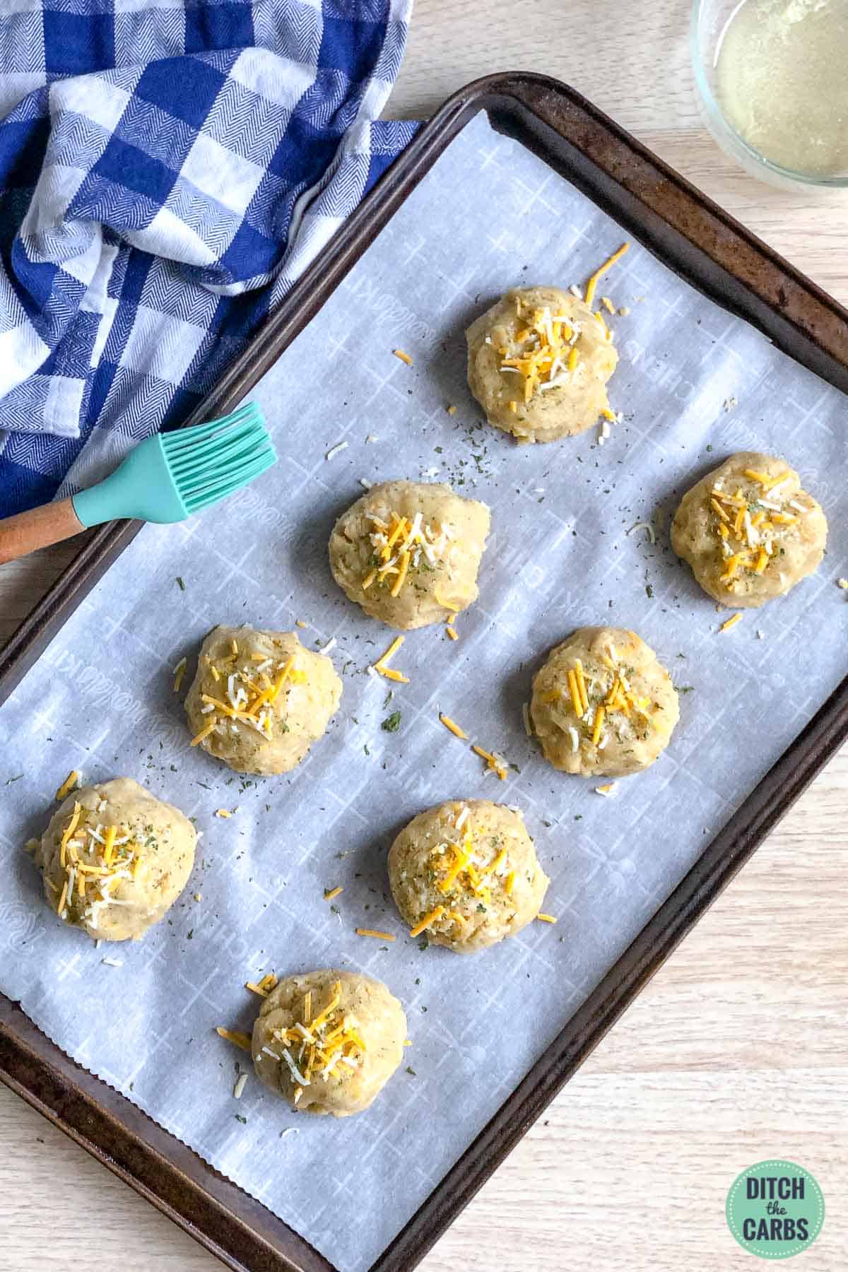 Keto cheddar biscuit(scone) dough has been arrange into 8 mounts on a lined baking pan. They have been brushed with melted butter and sprinkled with shredded cheese.
