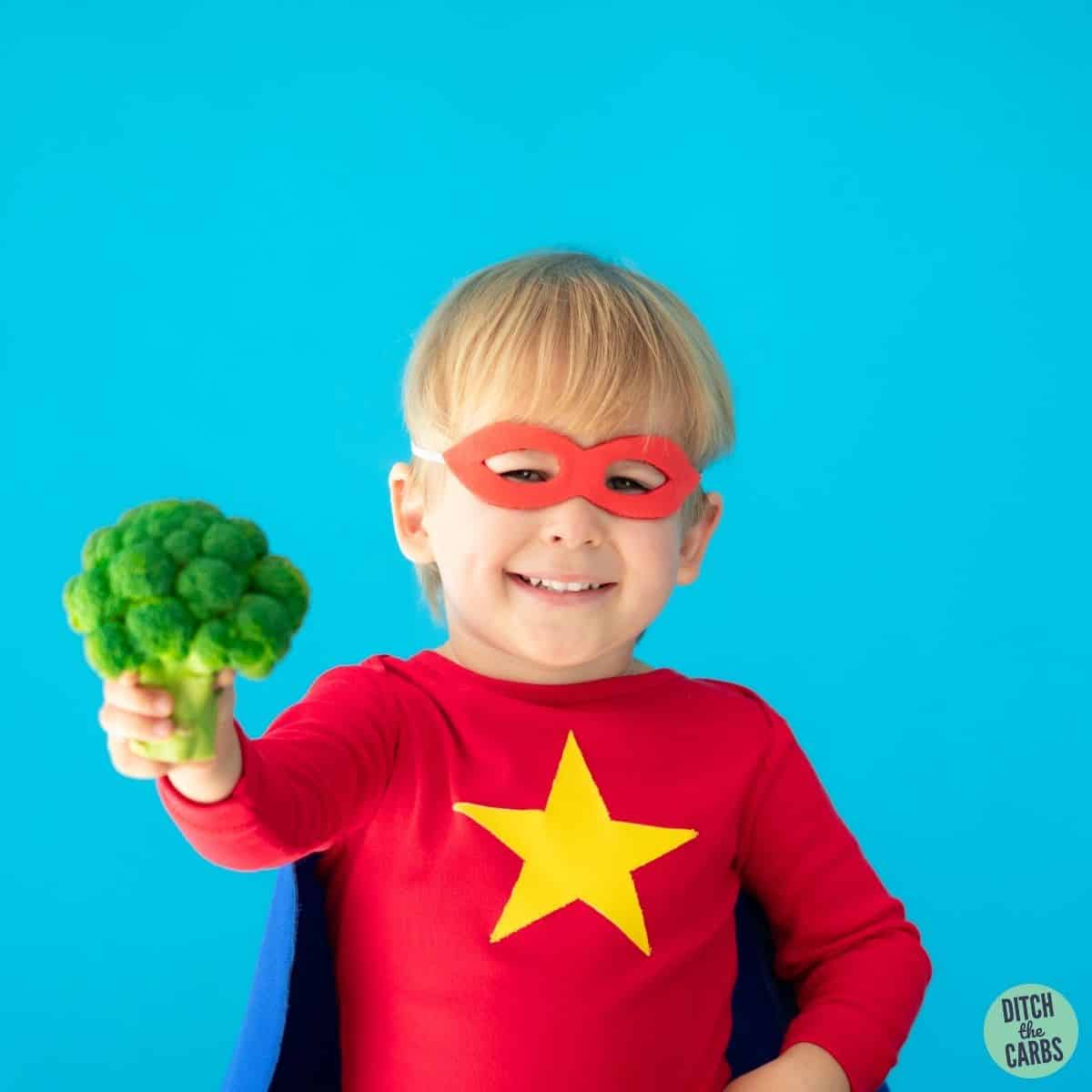a healthy child holding broccoli in a super hero outfit
