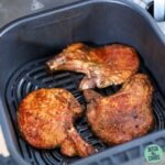 The air fryer basket has just been opened to show 3 seasoned and cooked bone-in pork chops in the basket.