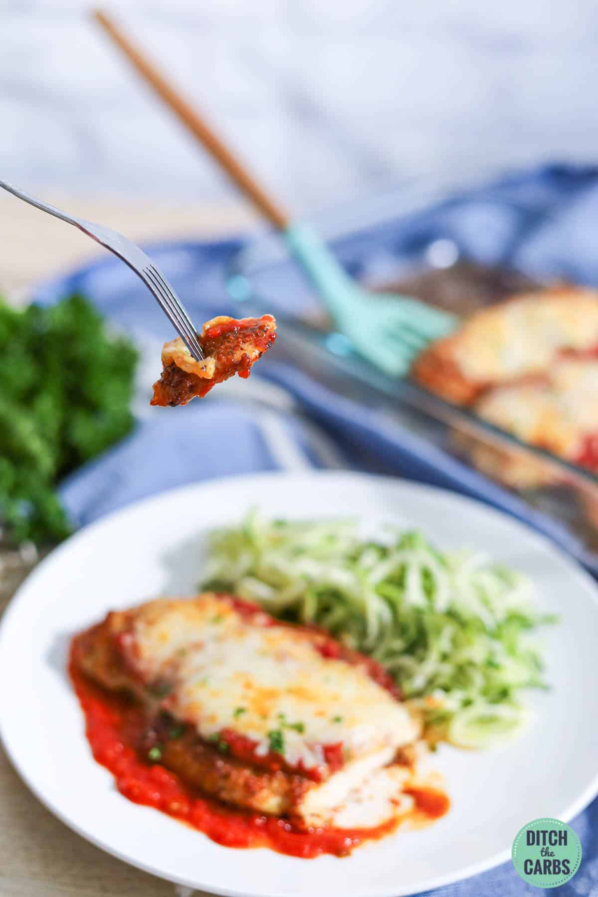 A fork is lifting a bite of keto chicken parmesan from a plate full of zucchini noodles and chicken parmesan.