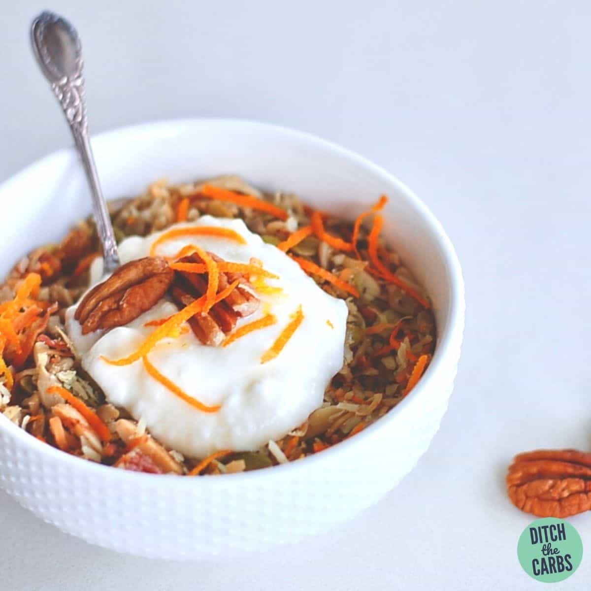 A bowl of gluten-free granola garnished with orange zest and pecans served with a silver spoon and yoghurt