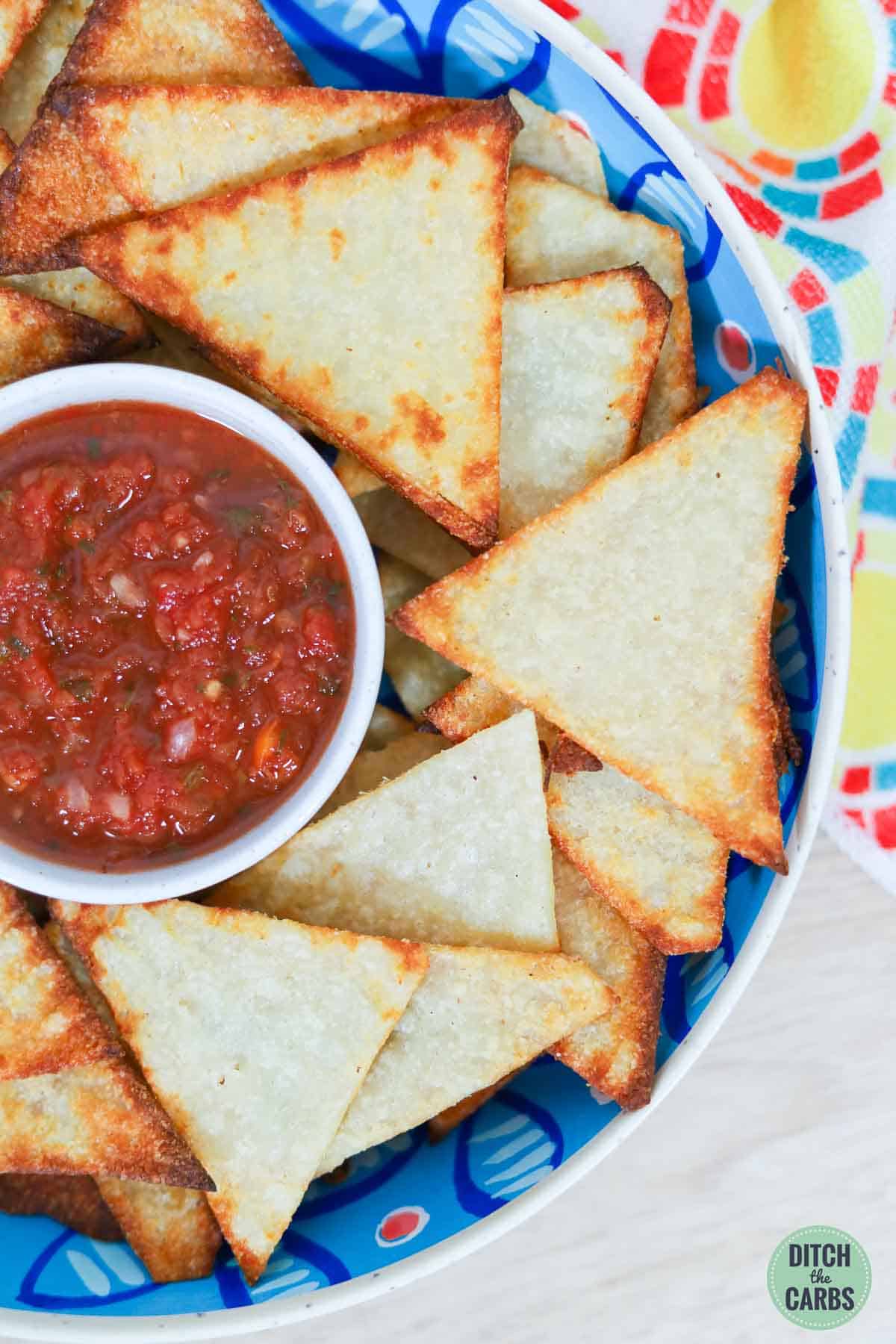 Close up overhead image of keto tortilla chips in a serving bowl with a dish of fresh salsa in the middle.
