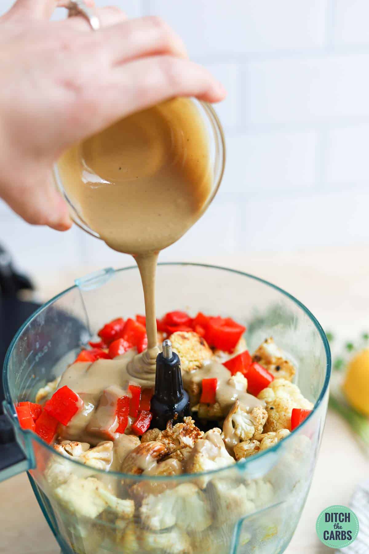 Pouring tahini over the roasted cauliflower and red peppers in the food processor.