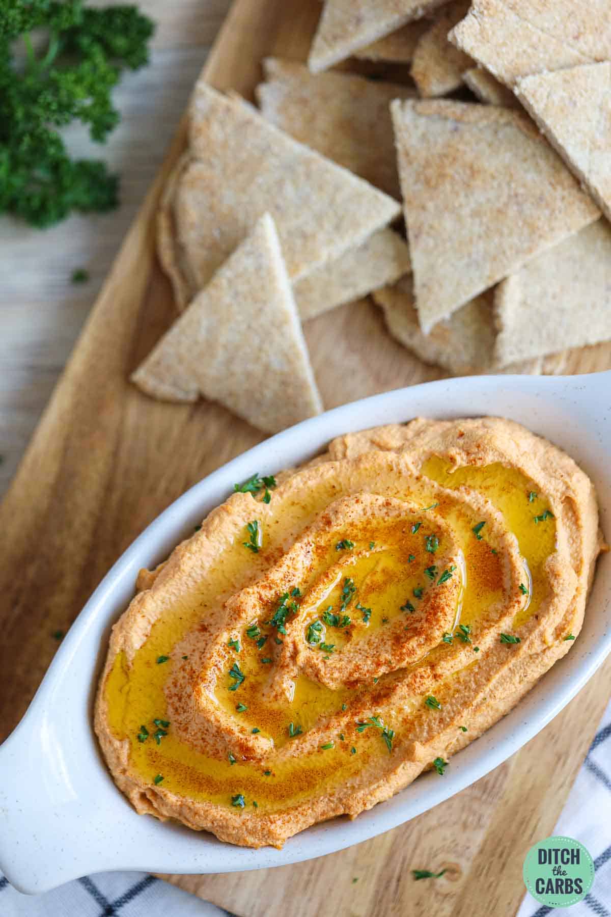 Roasted cauliflower low-carb hummus in a serving dish topped with parsley. It is resting a wooden tray with low-carb pita bread in the back for dipping.