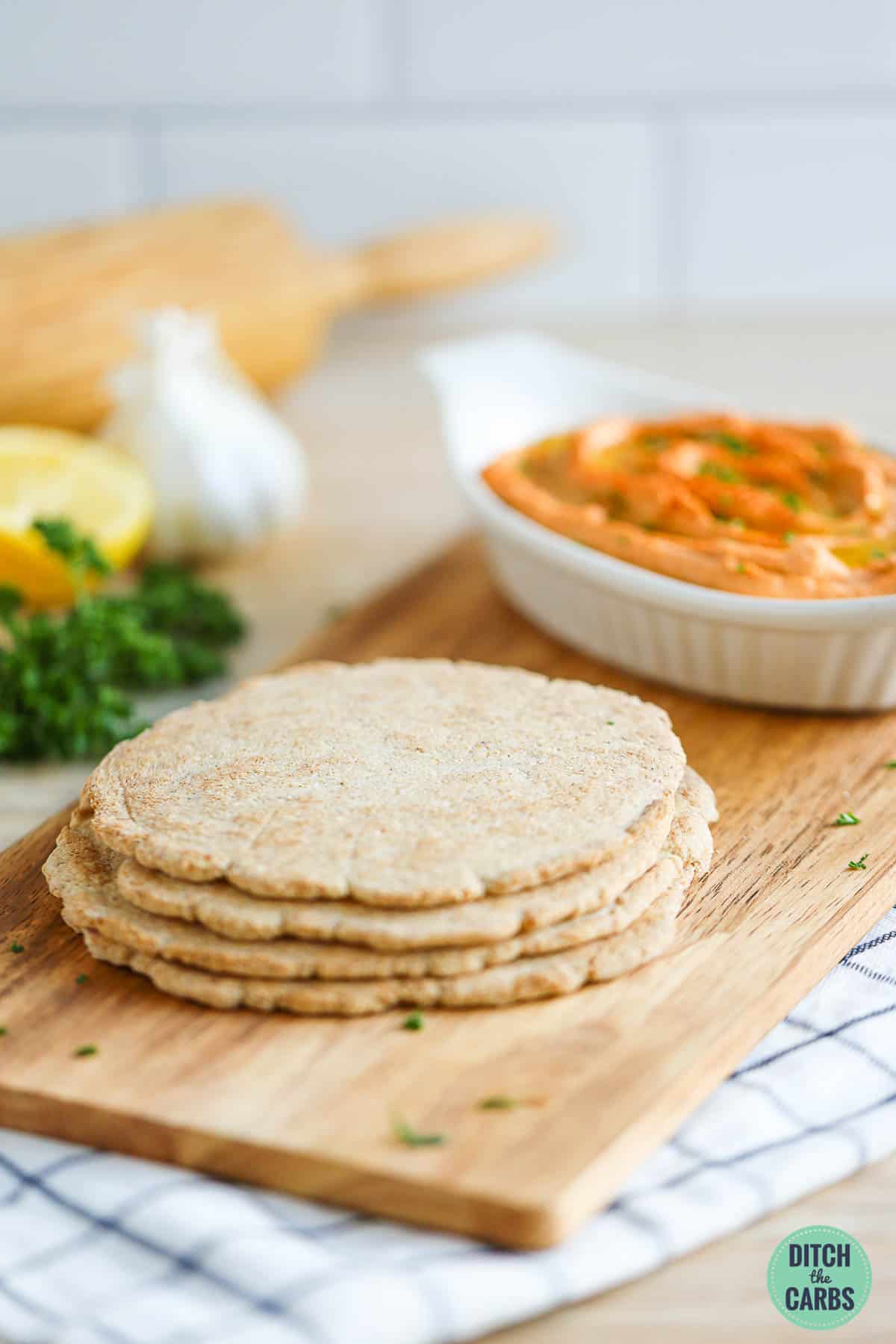 Low-carb pita bread baked and stacked on top of each other on a wooden tray. A dish of keto hummus is in the background.