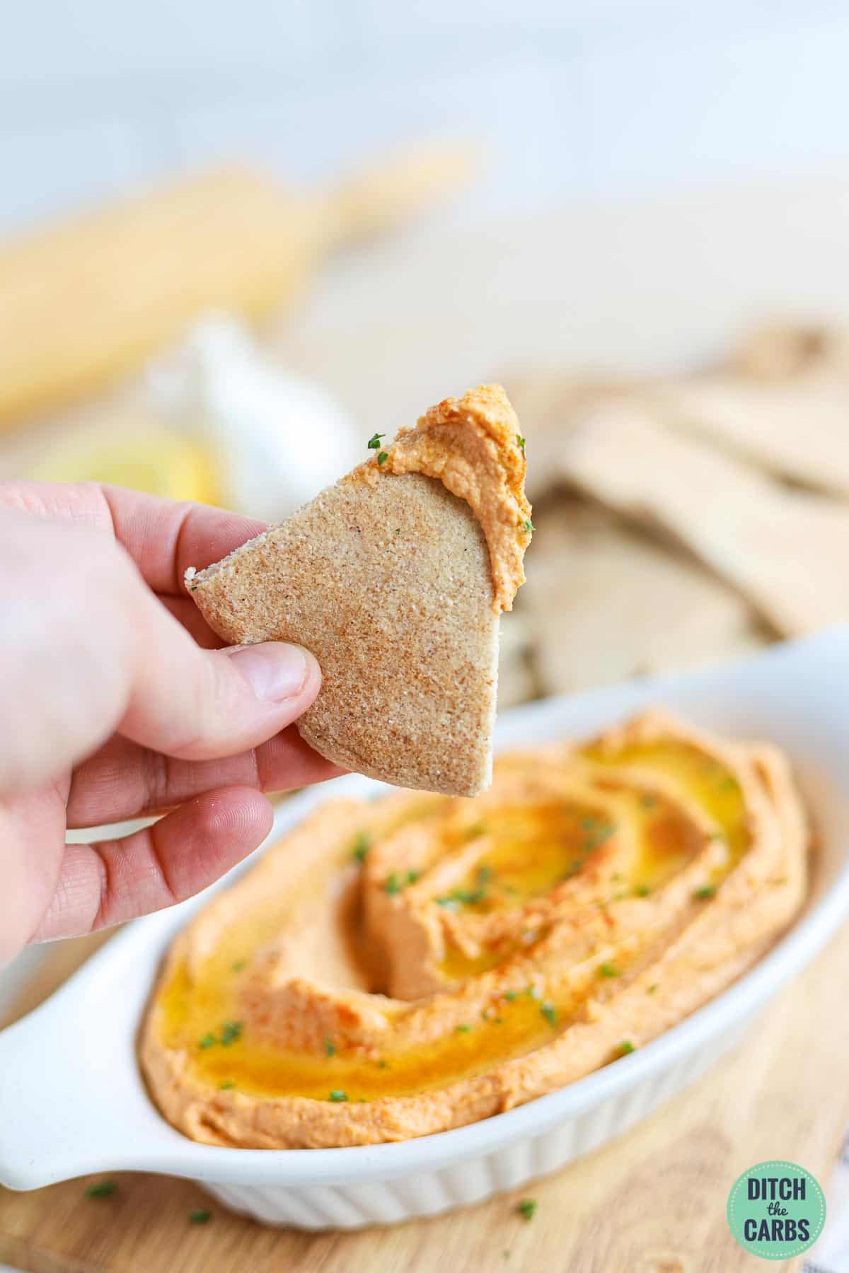 A hand is lifting a low-carb pita cut into triangle up from a dish of hummus.